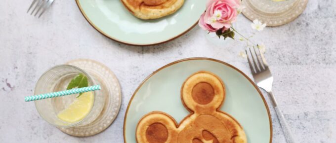 Two mickey mouse pancakes on different plates with drinks and forks on the side.