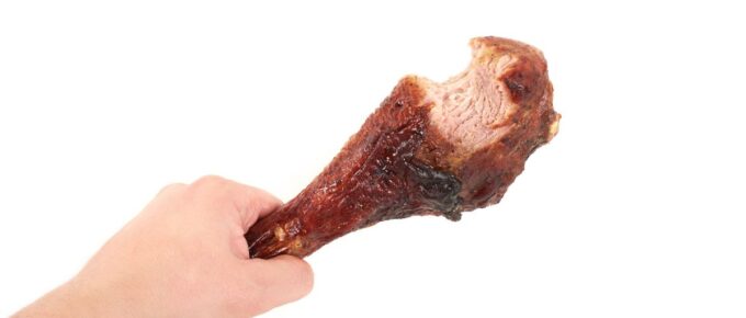 Hand holding roasted chicken leg with bite taken, isolated on white background.